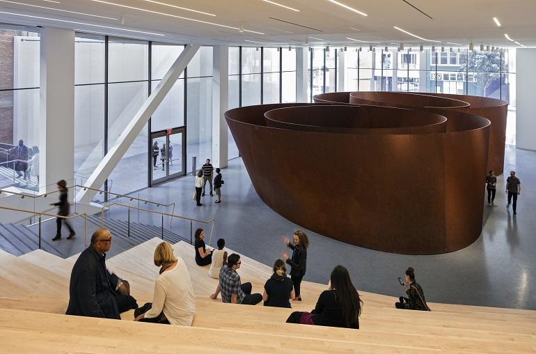 Large bronze sculptural installation in modern museum gallery with visitors and floor-to-ceiling windows overlooking city street.