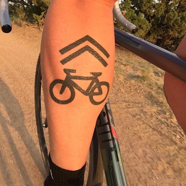 Cyclist's leg with sharrow tattoo on calf, holding bicycle handlebar.
