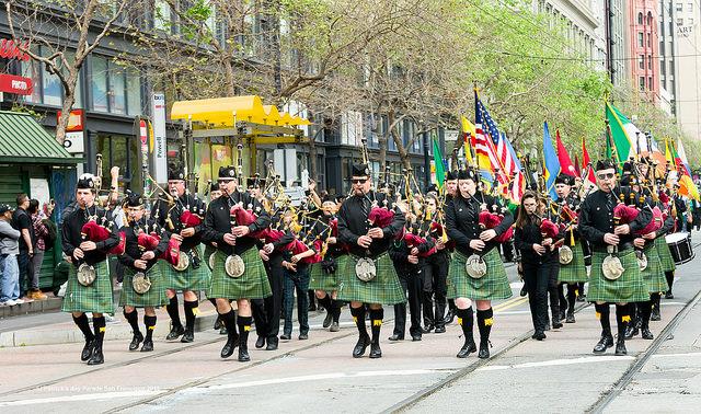 Bagpipe players in green kilts marching down a city street during a parade with international flags visible.