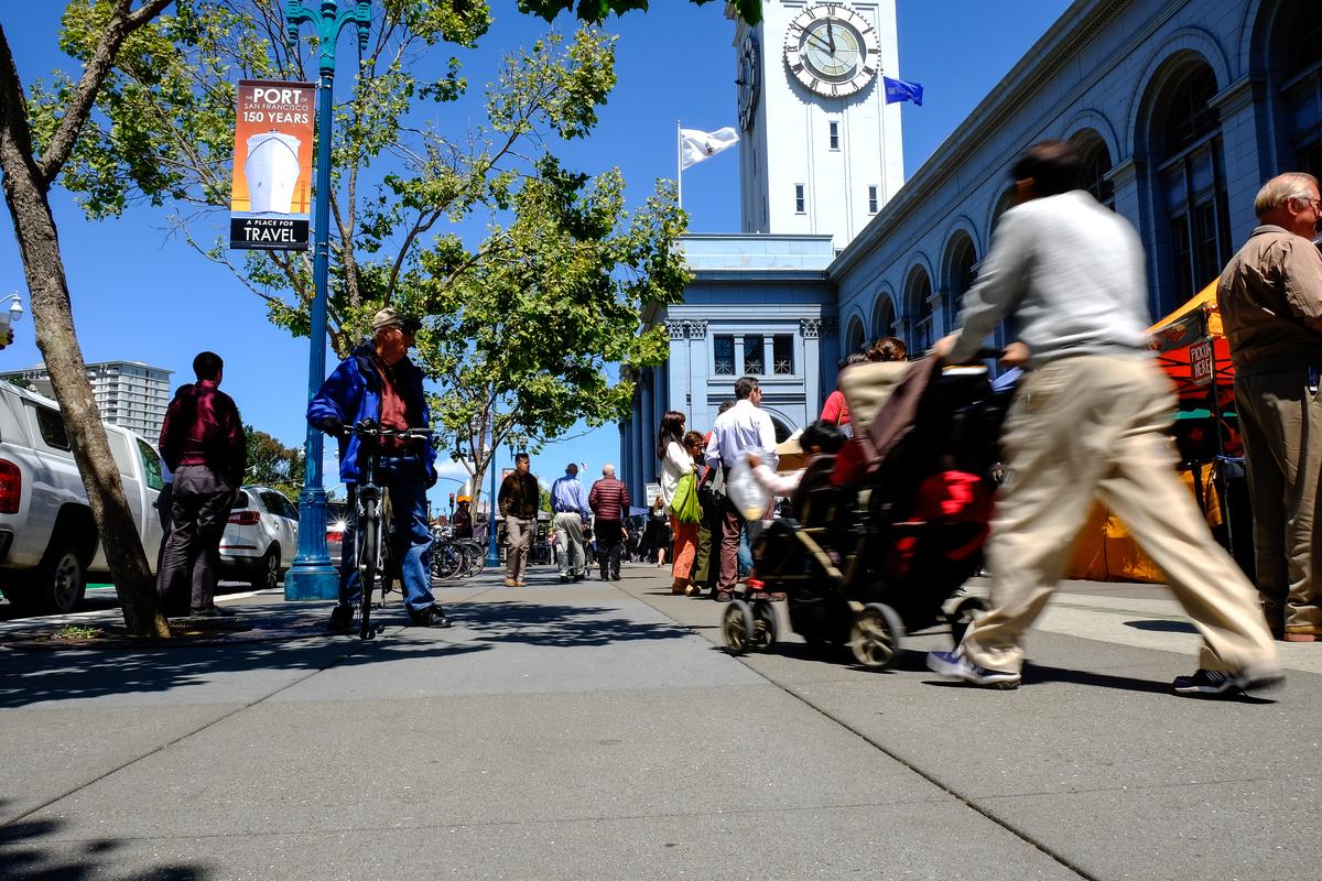 Pedestrians, cyclists, and strollers on tree-lined Embarcadero street with Ferry Building clock tower in background on sunny day.