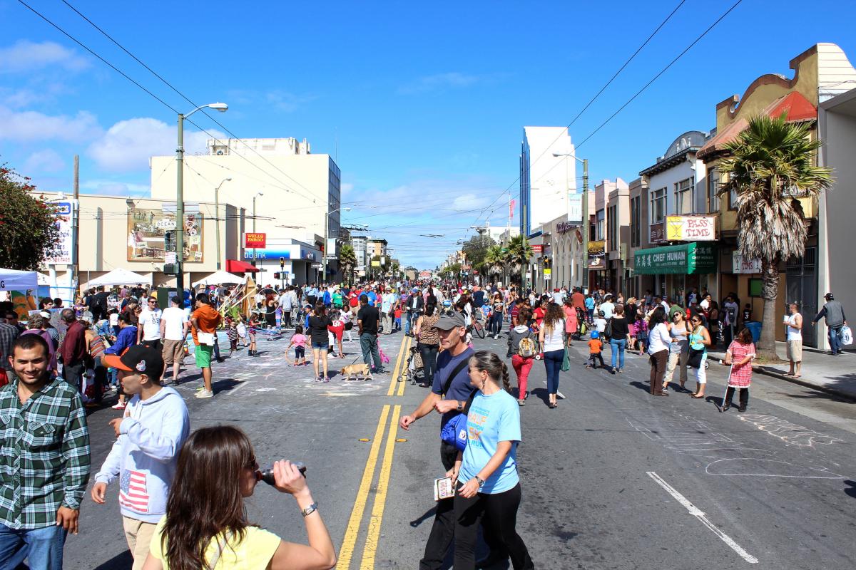 Crowded street festival with hundreds of pedestrians, vendor tents, and storefronts along Mission Street on a sunny day.