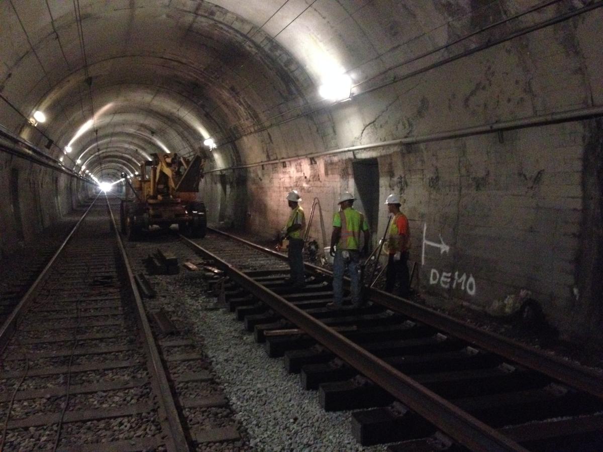 Workers in hard hats position rail on new ties inside an illuminated tunnel during track replacement work.