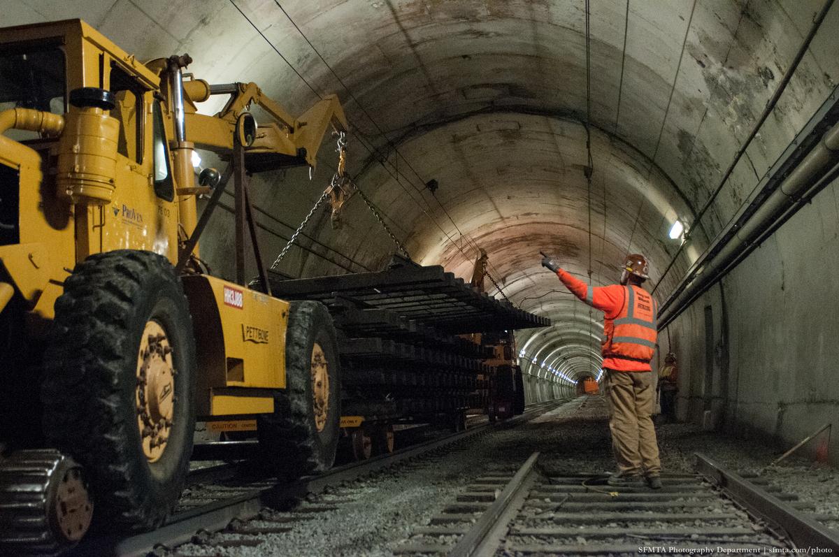 Yellow loader with chain hoist lowers steel beam in concrete tunnel while worker in orange vest directs from track below.