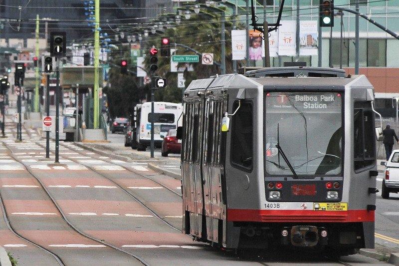 Light rail vehicle stopped at intersection on 3rd Street with traffic signals and street infrastructure visible.