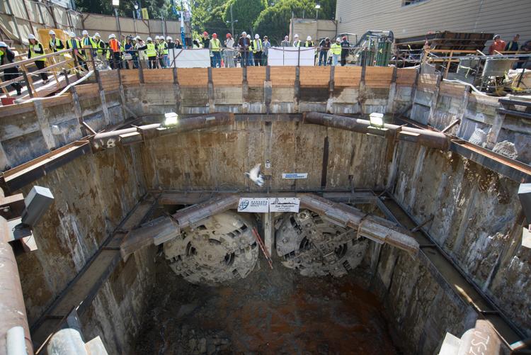 Large underground shaft with two tunnel boring machine cutter heads visible at bottom; workers and observers line the top perimeter.