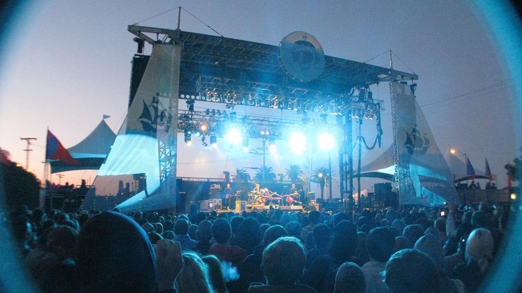 Large outdoor music festival stage with crowd at dusk, blue lighting illuminating performers and audience.