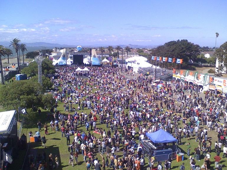 Aerial view of thousands of people gathered on a grassy field at an outdoor music festival with stage and tents visible.