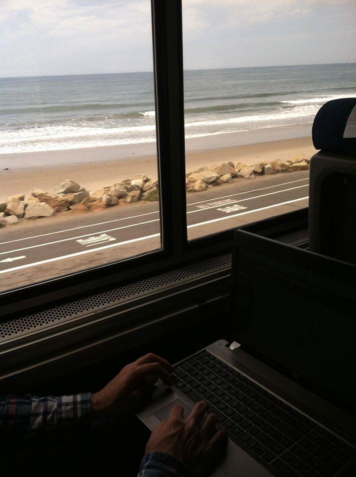 Person working on laptop aboard Amtrak Coast Starlight train with Pacific Ocean and beach visible through window.