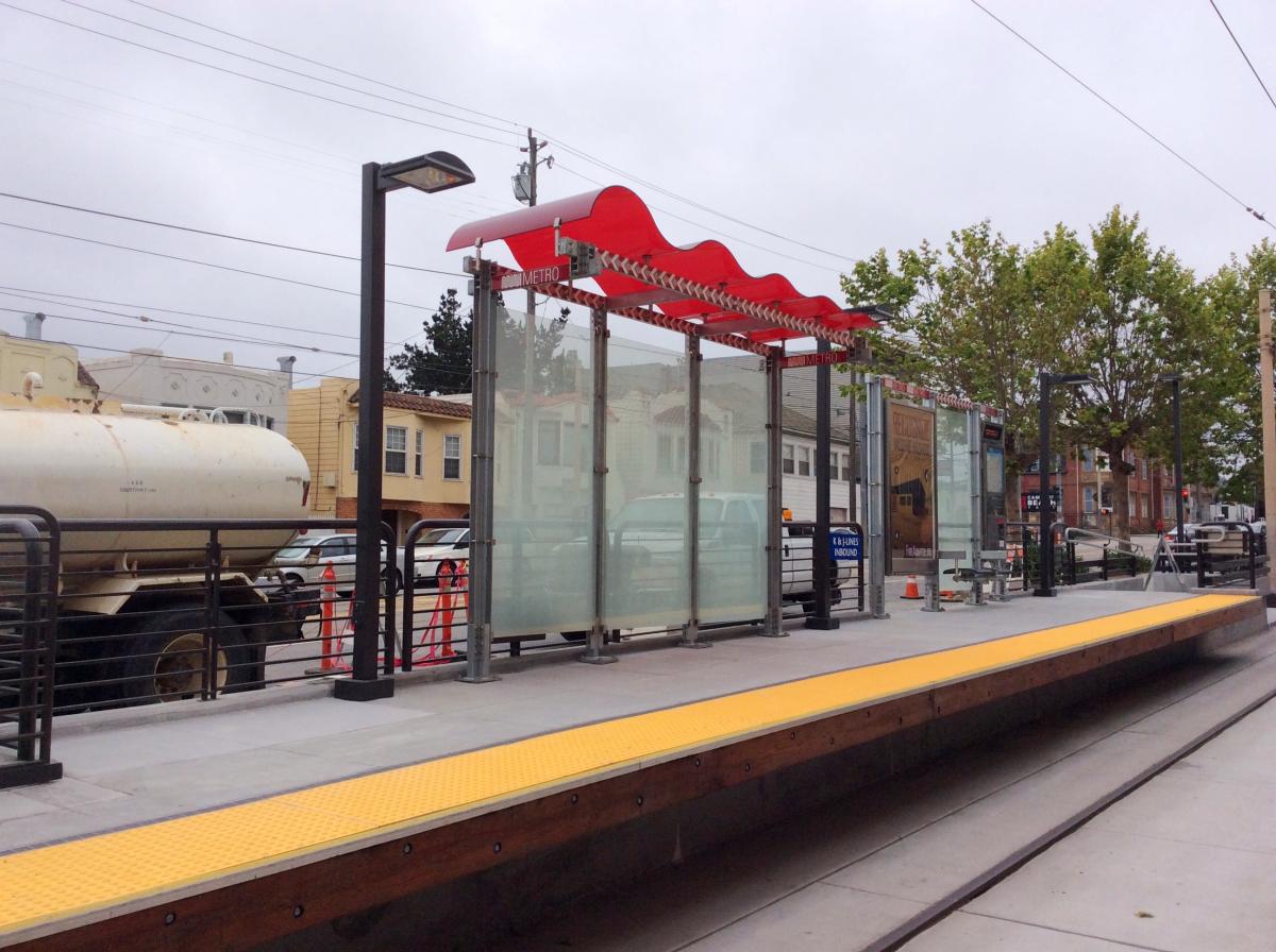 Elevated transit platform with red wave-shaped shelter, glass panels, and yellow tactile warning strip along track.