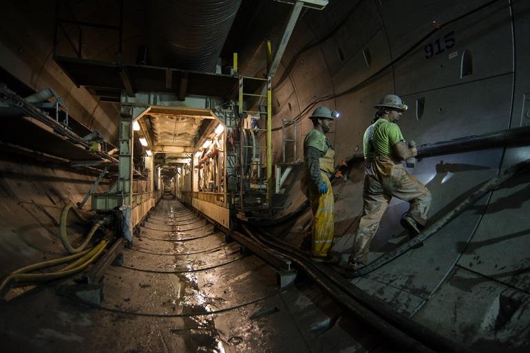 Workers in hard hats and safety gear operate equipment inside an underground transit tunnel under construction.