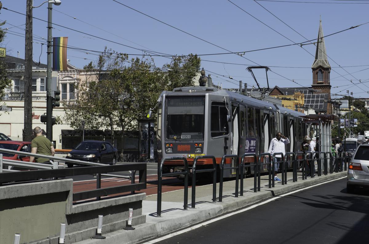 Light rail vehicle stopped at platform with passengers boarding at street-level station surrounded by urban buildings and infrastructure.