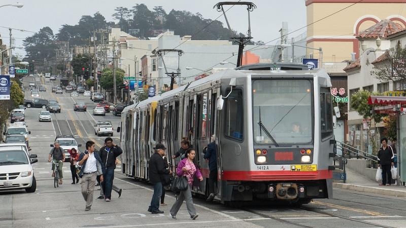 Light rail train stopped on Taraval Street with passengers boarding and alighting in the roadway.
