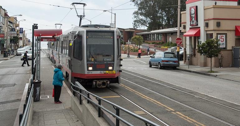 Red and white light rail vehicle stopped at boarding platform with passenger waiting on sidewalk.