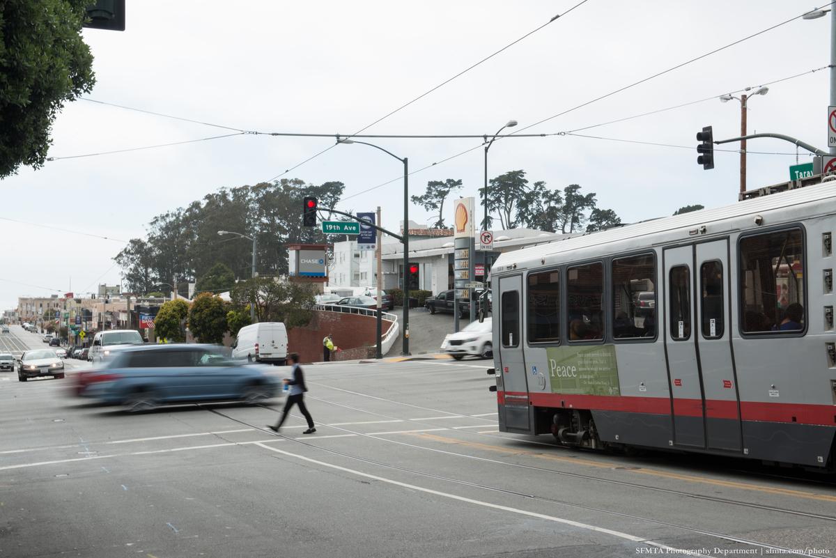 Light rail vehicle at busy urban intersection with pedestrian crossing and vehicle traffic.