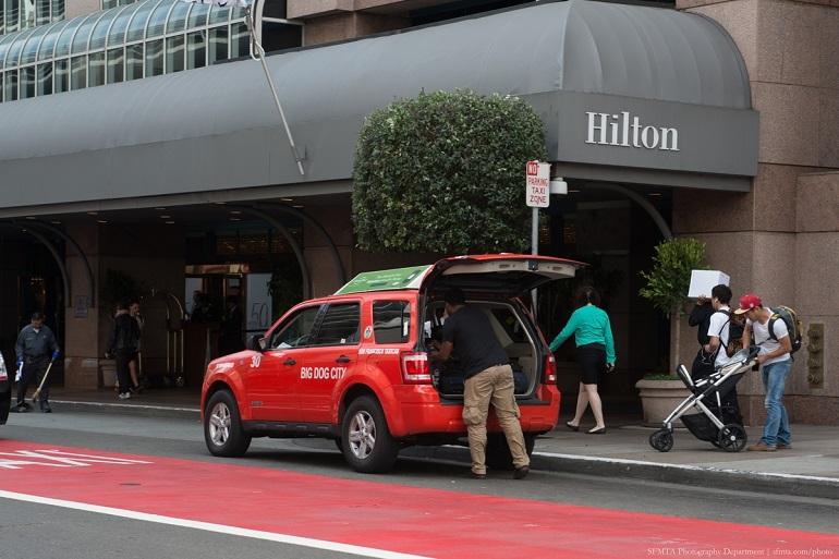 Red taxi parked at Hilton hotel entrance with driver loading luggage and pedestrians on sidewalk.