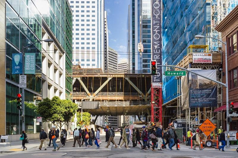 Crowded crosswalk at Fremont and Howard Streets with Transbay Transit Center under construction overhead.