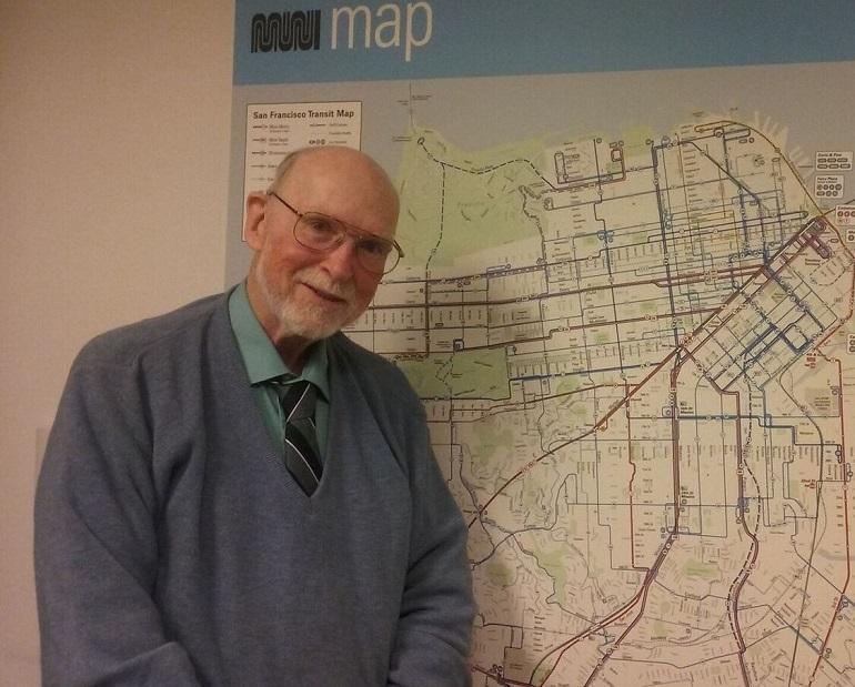 Elderly man with glasses smiling in front of a San Francisco Muni transit map on a wall.