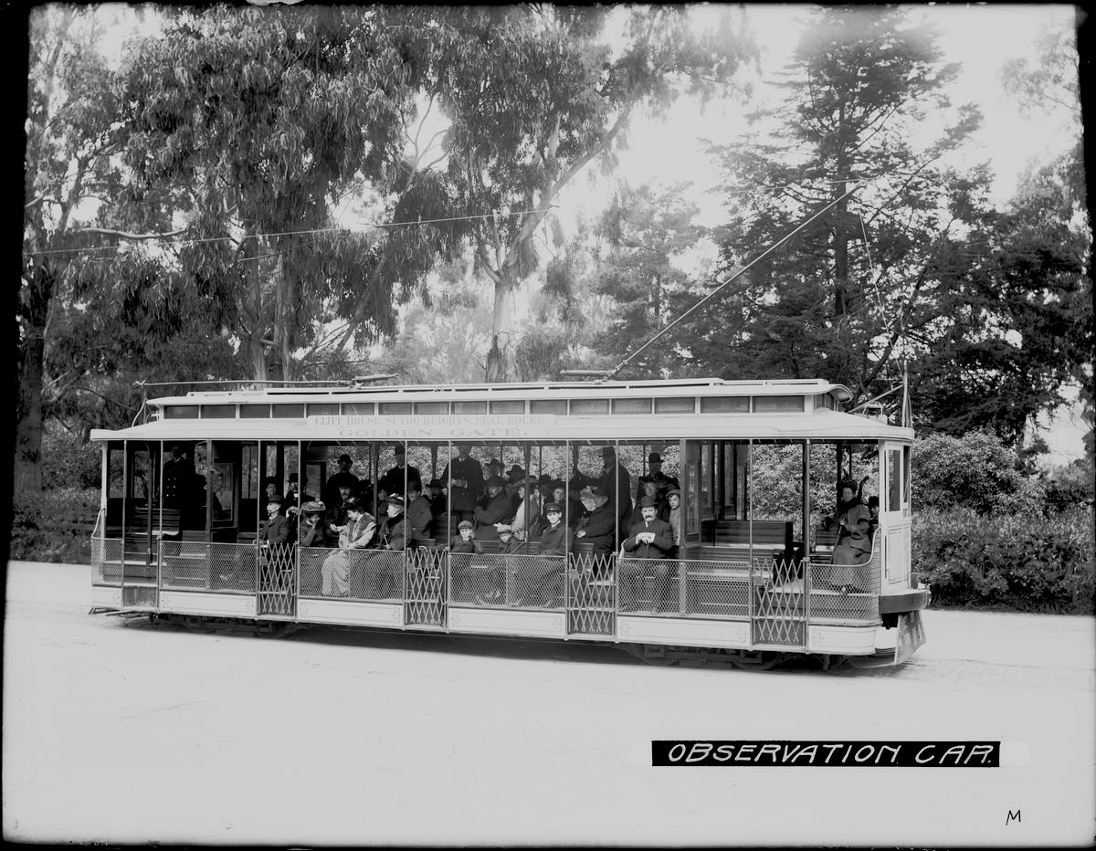 Open-air observation streetcar filled with passengers in a tree-lined park setting, 1904.