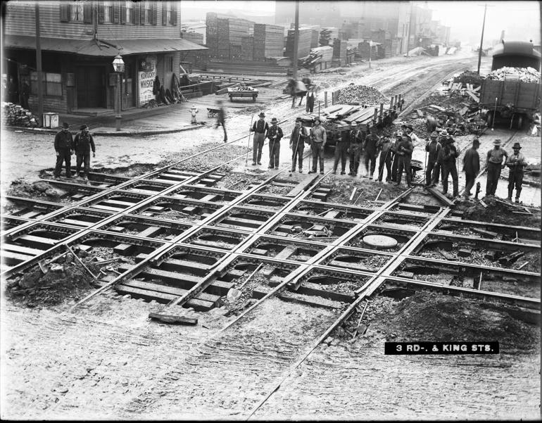 Workers stand beside newly laid railroad tracks and streetcar crossing at 3rd and King Streets, 1904.