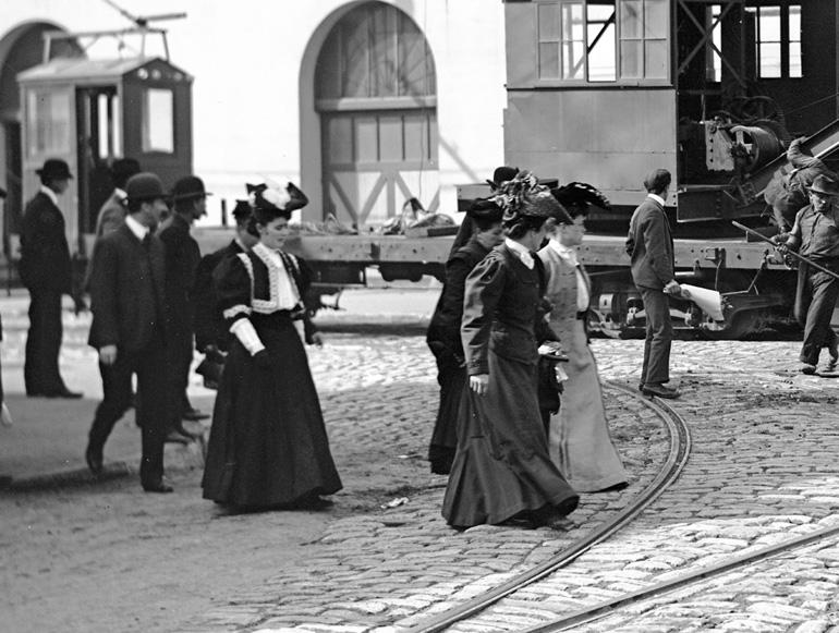 Women in Victorian-era clothing walk across cobblestone street near cable car tracks, 1905.