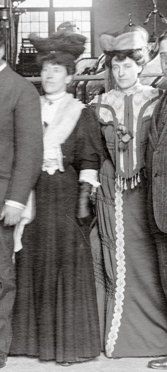 Two women in Edwardian-era formal attire with large hats stand indoors near a window.
