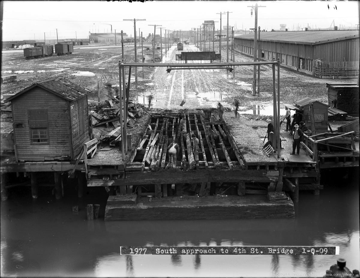 Southern approach to 4th Street Bridge showing muddy terrain, wooden pilings, industrial structures, and waterfront activity, 1909.