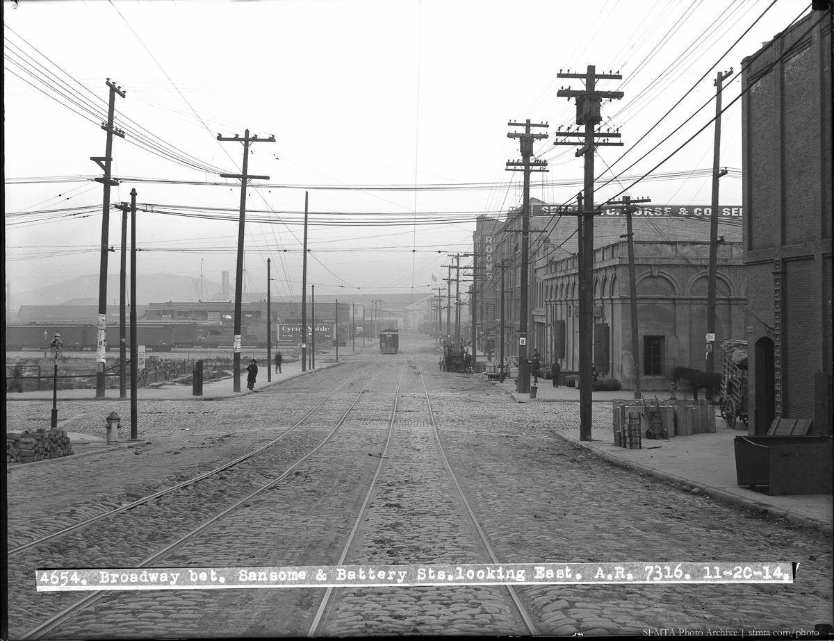 Broadway and Battery Street looking east, 1914. Streetcar tracks, wooden utility poles, brick buildings, and unpaved street.