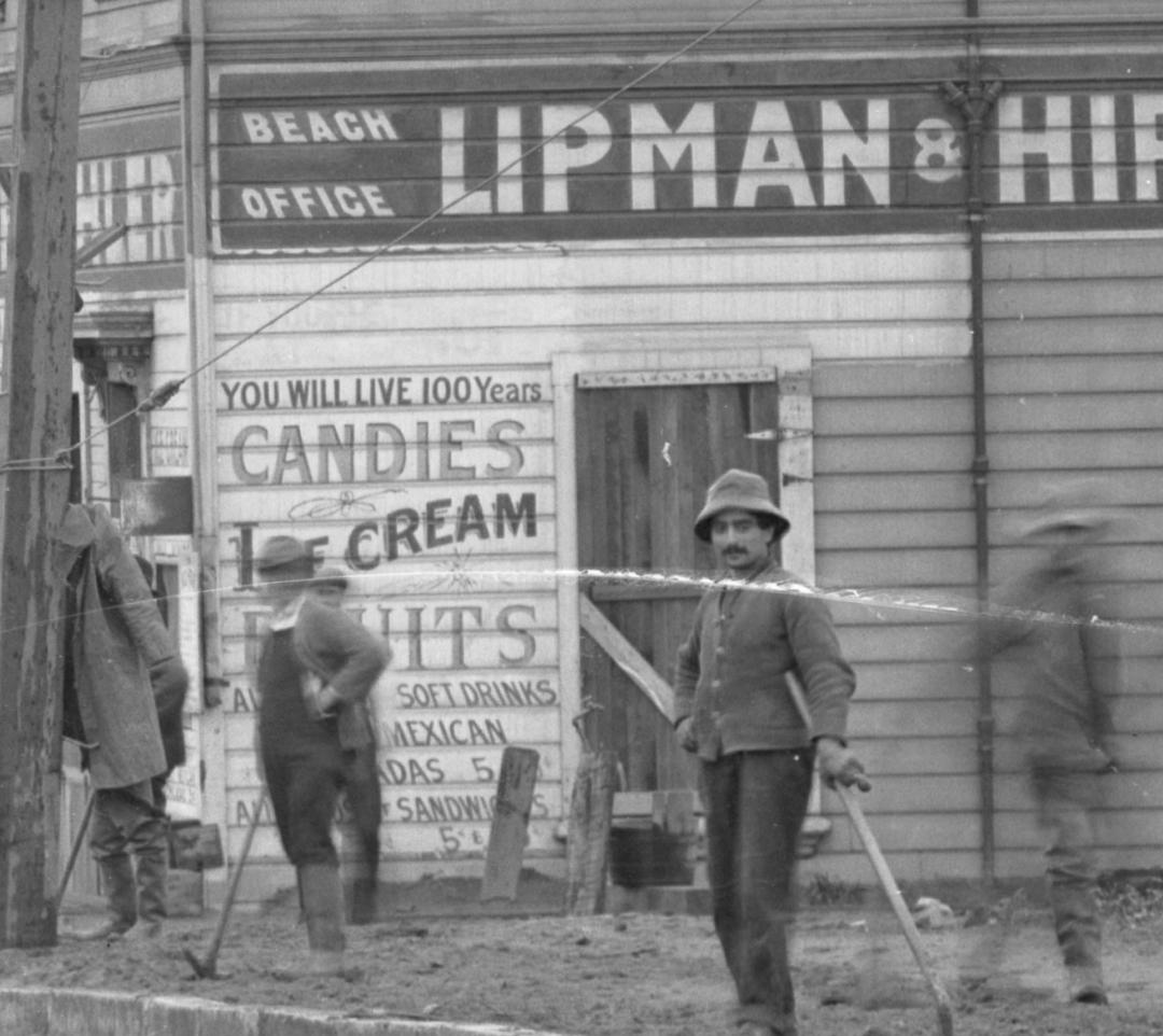Black and white photograph of workers on a beach with vintage storefront signs advertising candies, ice cream, and soft drinks, circa 1915.