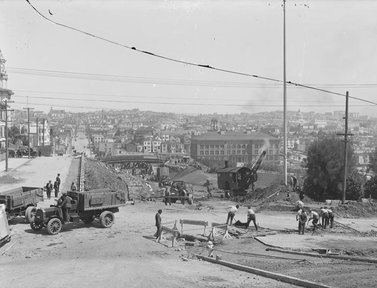 Construction site with trucks and workers at 20th and Church Streets, 1916, with Dolores Park and city skyline visible.