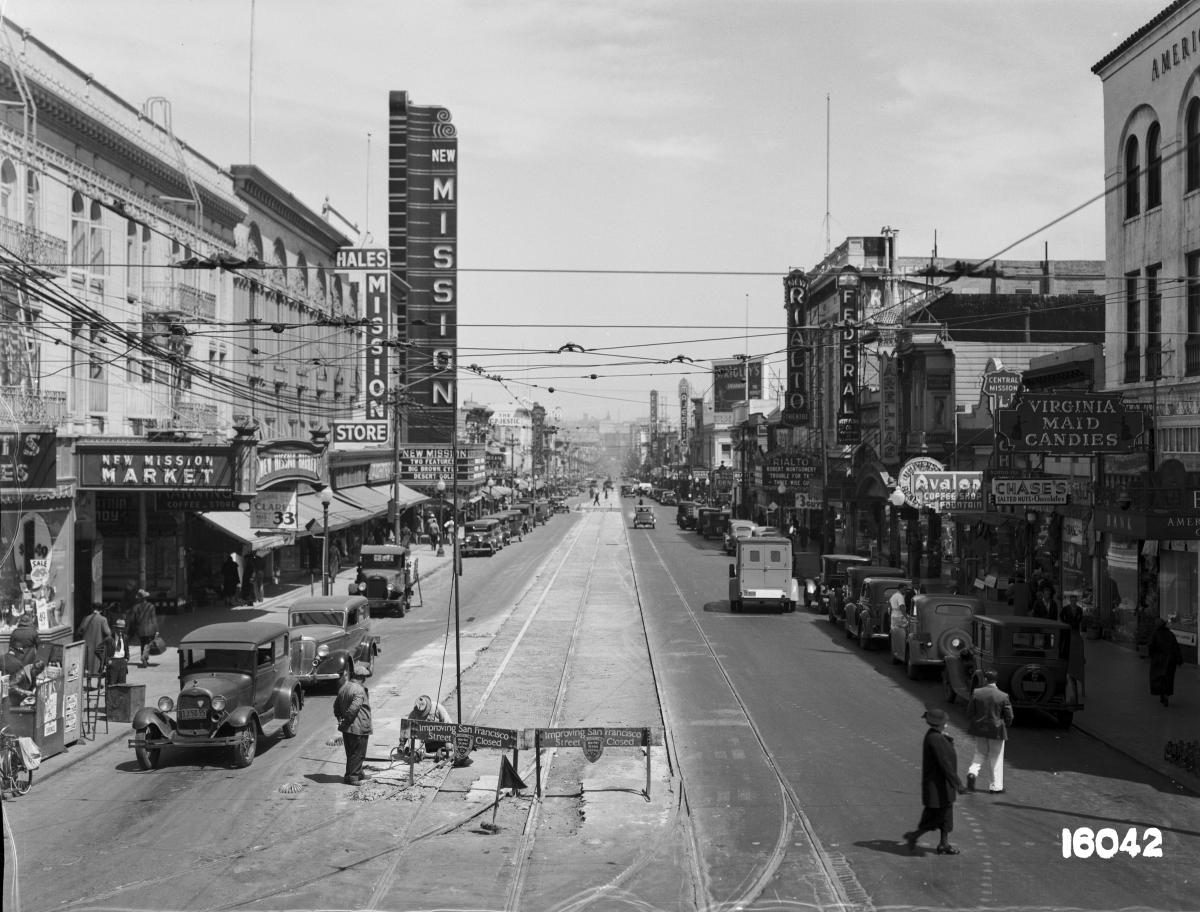 Mission Street looking north with streetcar tracks, storefronts, and vintage vehicles, circa 1936.