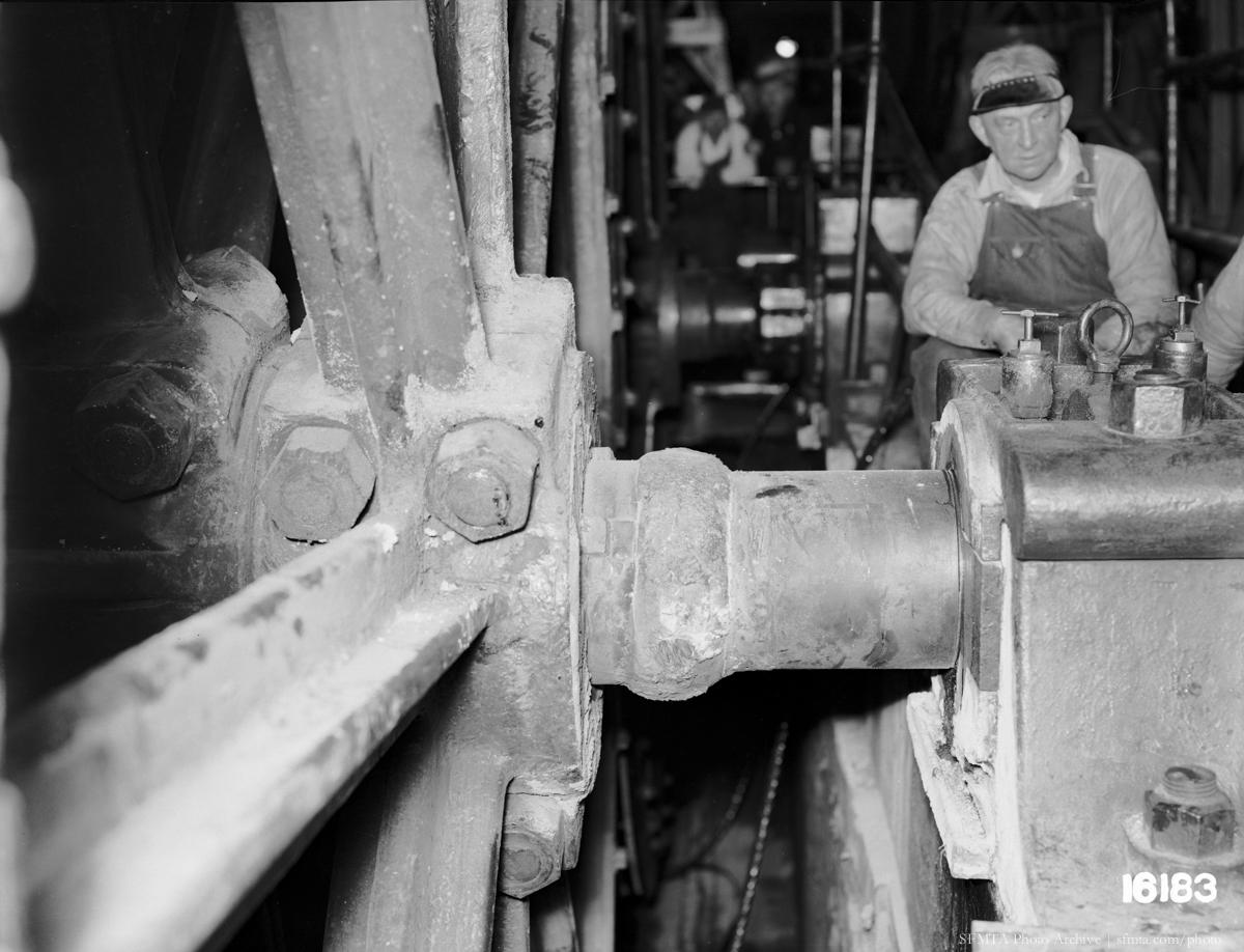 Black and white photograph of a welded shaft with a worker in the background at an industrial powerhouse facility.