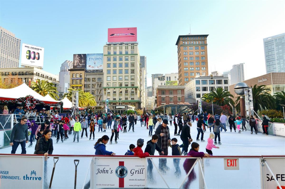 Crowds of people ice skating at Union Square with downtown San Francisco buildings in the background.