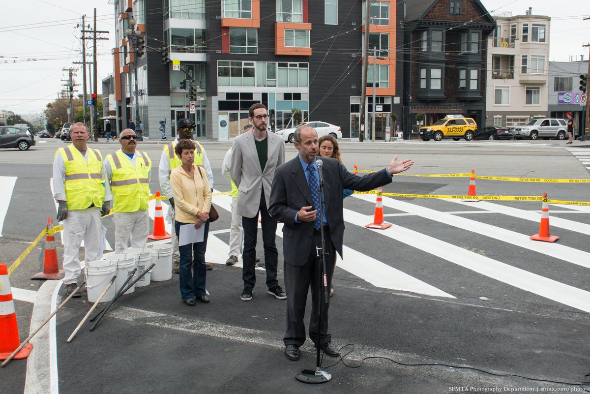 Man in dark suit speaks at microphone on Upper Market Street with safety crew and officials nearby, yellow caution tape and traffic cones visible.