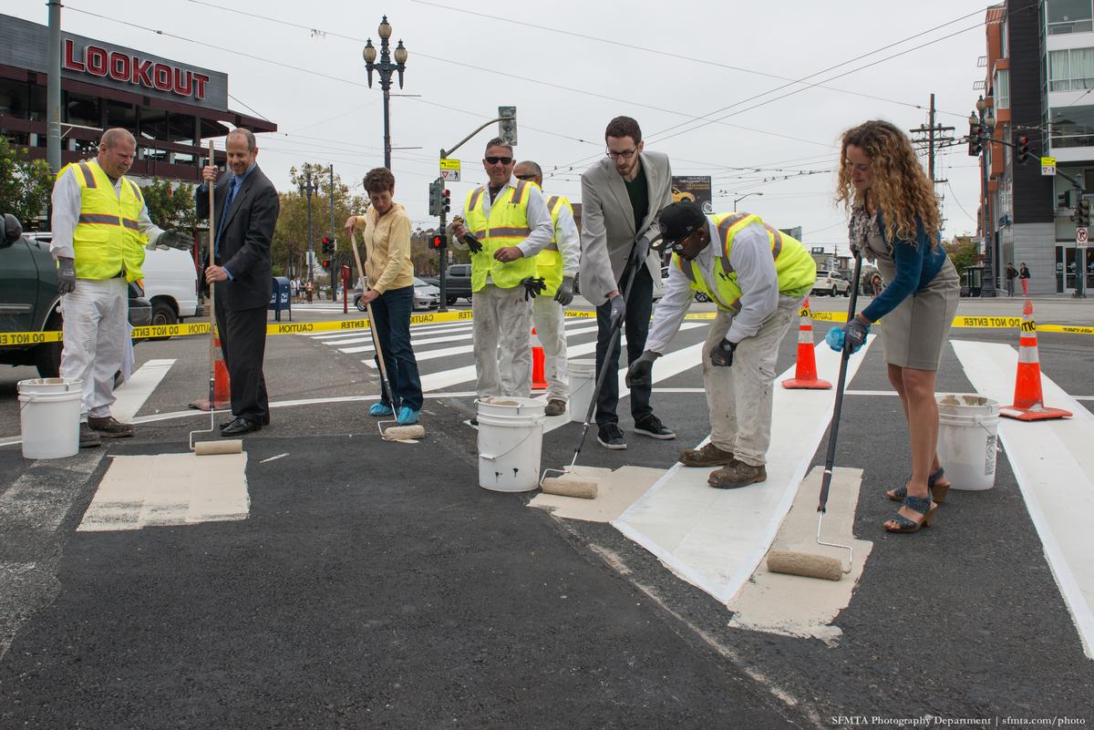 Group of workers and officials in safety vests painting a street safety zone with beige paint on a San Francisco street.