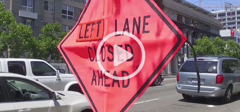 Pink diamond-shaped construction sign reading "LEFT LANE CLOSED AHEAD" on Van Ness Avenue with vehicles and buildings visible.