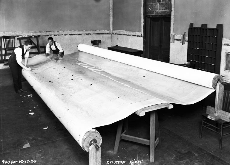 Workers unroll a large rolled map across the floor of an industrial room, circa 1923.