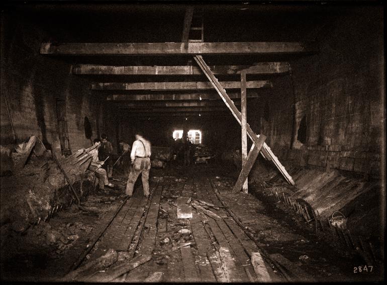Workers excavating tunnel interior with wooden support beams and rail tracks, circa 1916.