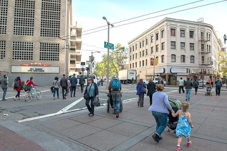 Pedestrians crossing streets at a busy urban intersection with historic buildings and storefronts.