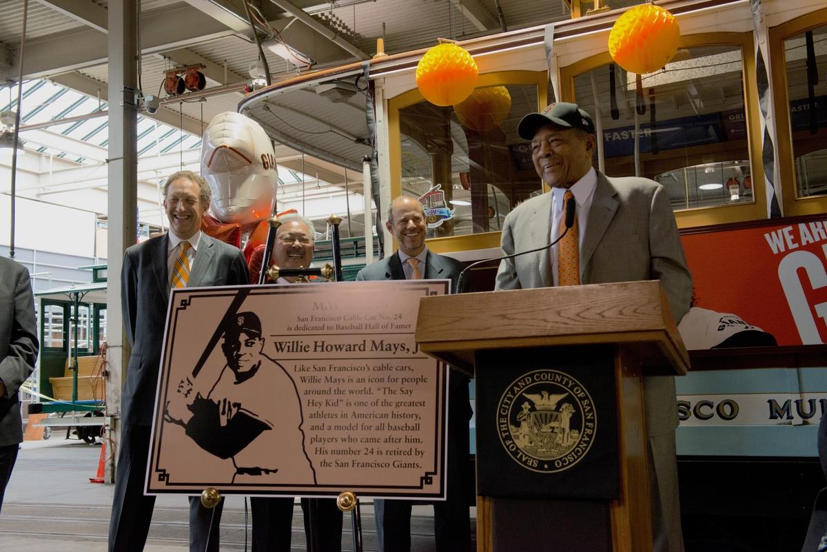 Four men stand at podium with Willie Mays plaque; decorated cable car behind them with yellow balloons.