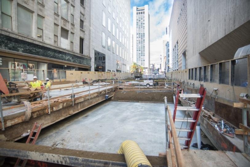 Construction site between downtown buildings with workers in safety vests preparing concrete foundation for temporary plaza.