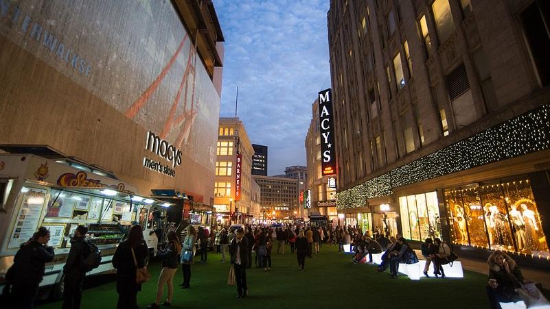 Crowded pedestrian plaza at night with Macy's storefront and illuminated buildings, people walking and gathering on green ground.
