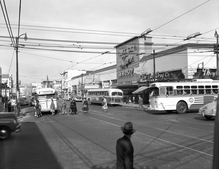 Busy street intersection with pedestrians, storefronts, and Muni motor coaches at Mission and 16th, 1949.