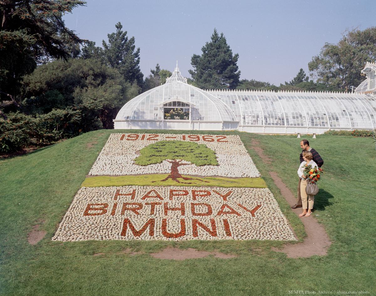 Floral plaque display celebrating Muni's 50th anniversary in 1962, with greenhouse and visitor in background.