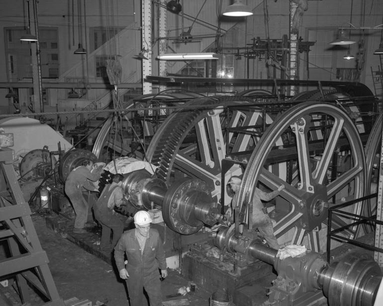 Workers lower a large geared driveshaft using a crane next to massive pulleys in an industrial workshop.