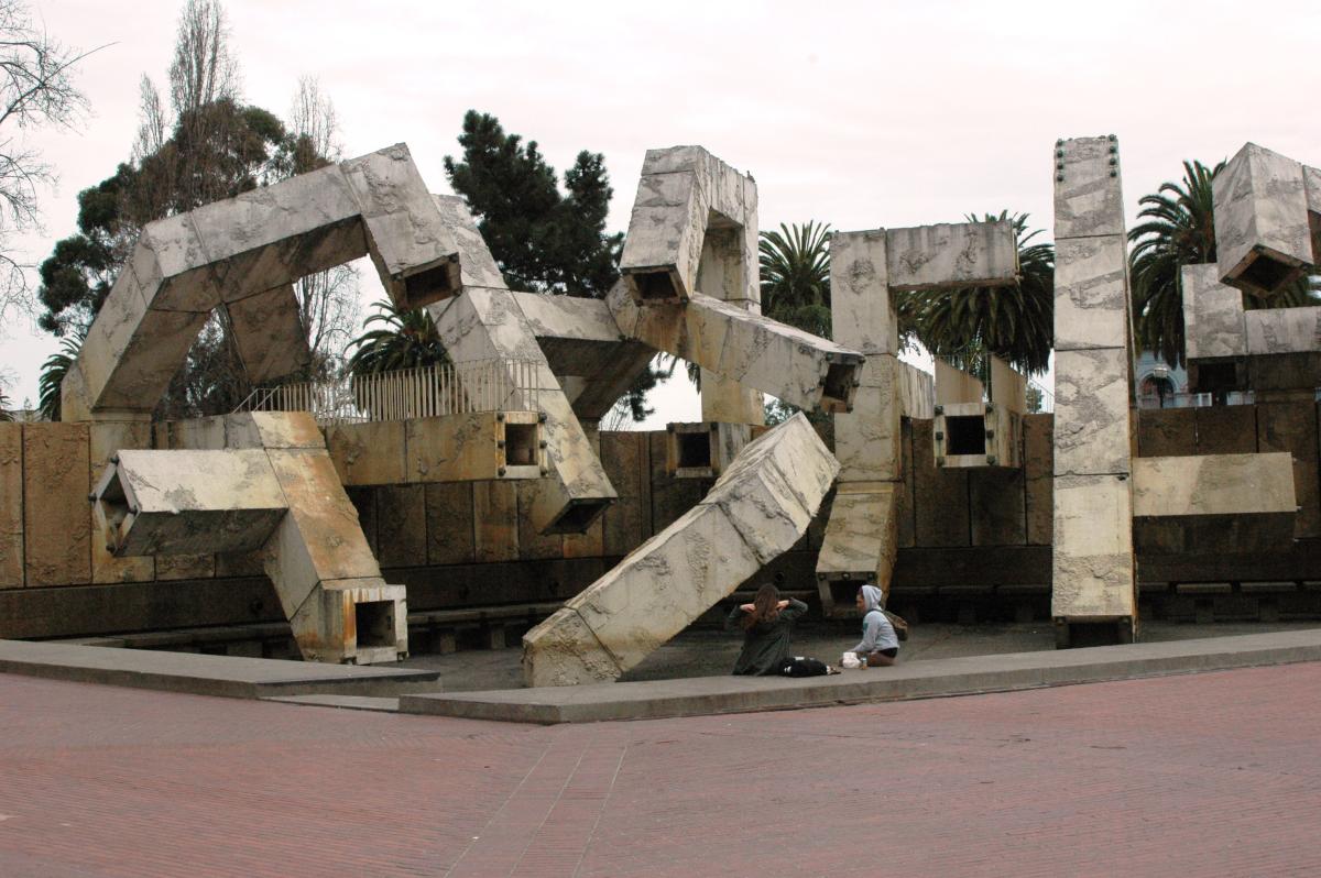 Vaillancourt Fountain concrete sculpture with angular blocks and palm trees, person sitting nearby on red brick plaza