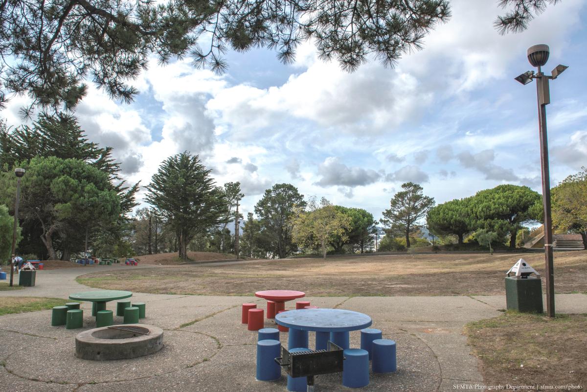 Hilltop Park picnic area with colorful picnic tables, fire pit, and open grassy field surrounded by trees.