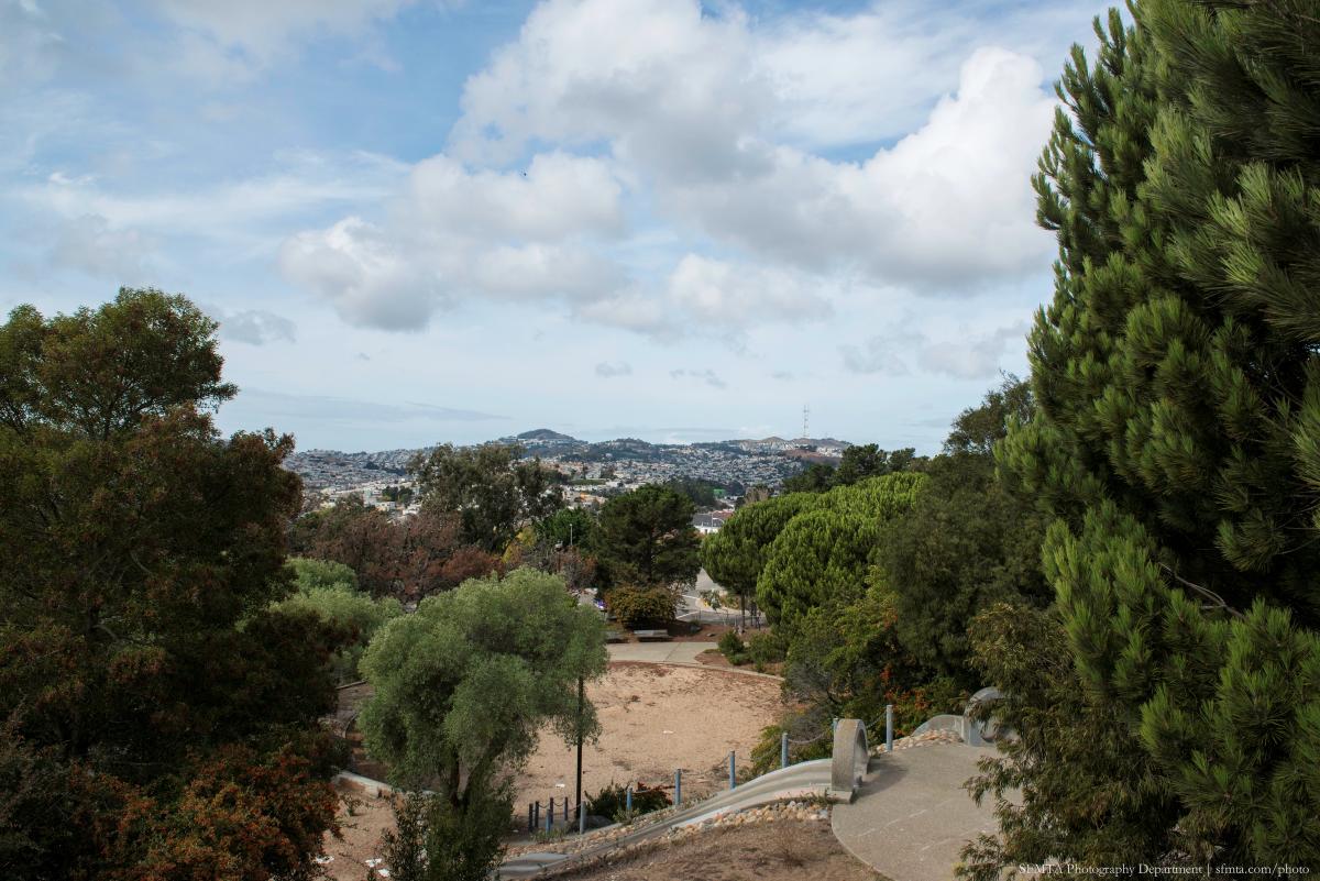 Hilltop Park picnic area with trees, open grounds, and distant city views under partly cloudy sky.