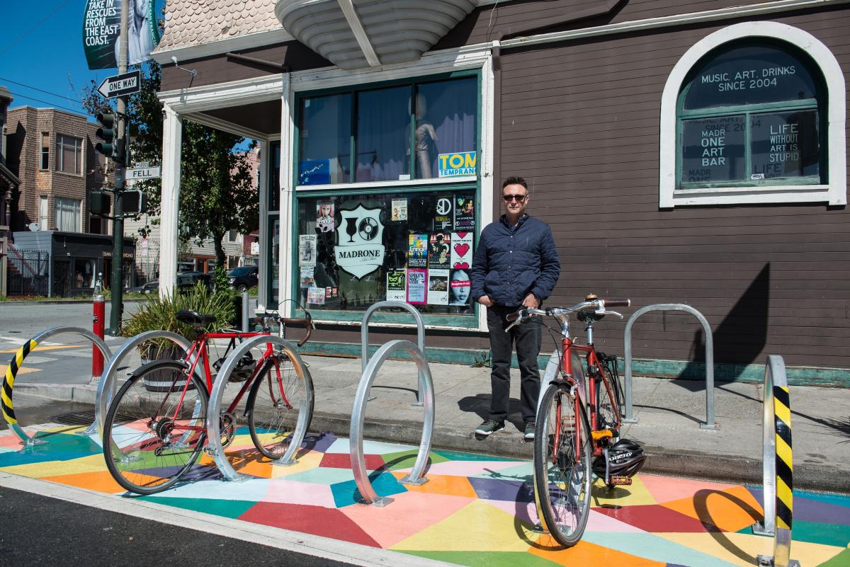 Man standing with red bicycle next to colorful bike racks in front of storefront with mural.