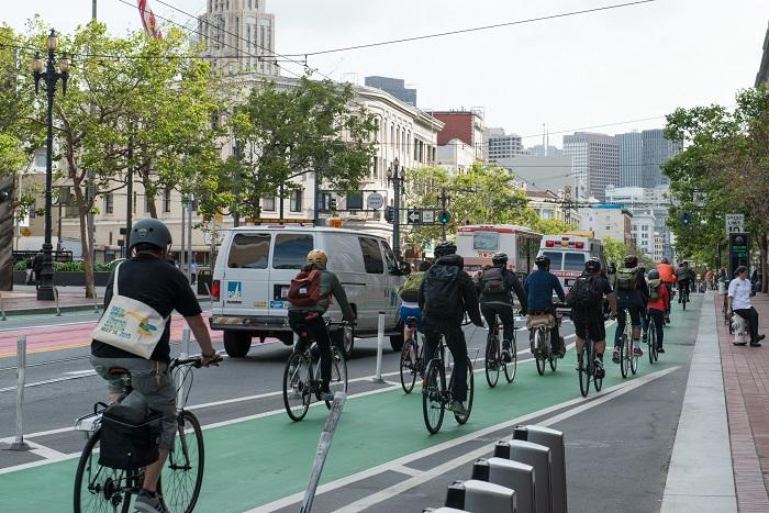 Group of cyclists riding in a protected green bike lane on a city street with buildings and trees in the background.