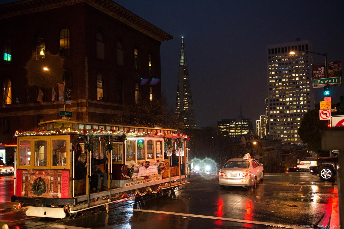 Decorated California Line cable car crosses Powell Street at night with downtown skyscrapers illuminated in background.
